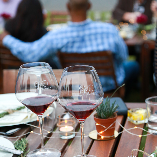 Two glasses of red wine on a table at Rendez-vous Winery, with guests enjoying a social gathering outdoors.