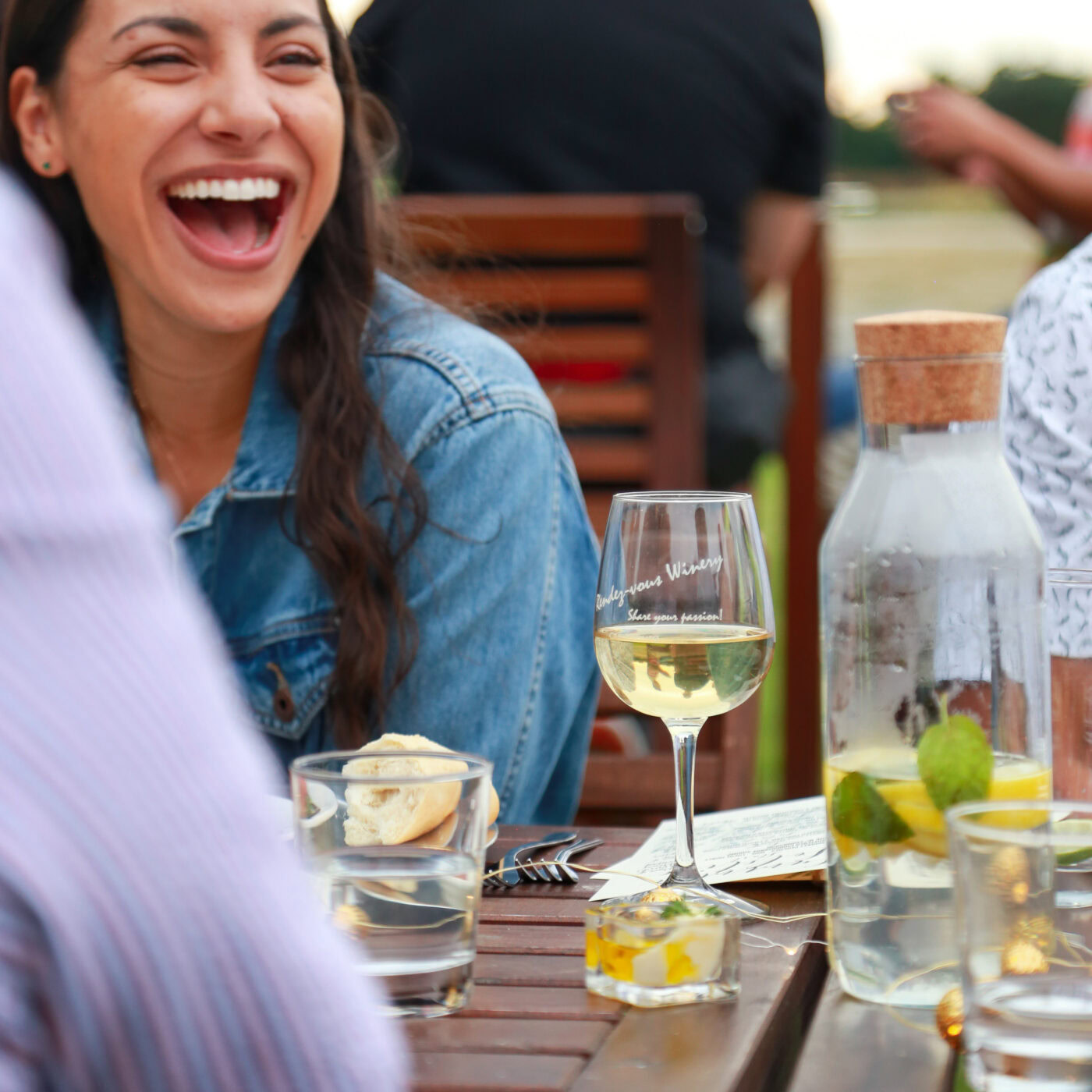 Woman laughing and enjoying wine outdoors at Rendez-vous Winery with friends, surrounded by glasses, water bottles, and a sce.